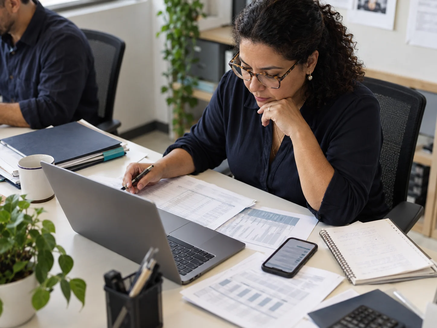 Profissional confere planilhas impressas ao lado do notebook, com anotações e papéis espalhados pela mesa.